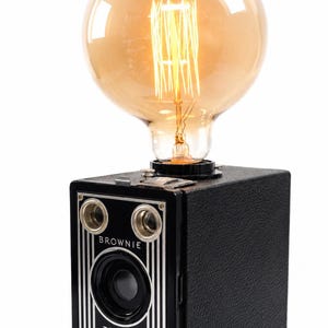 Könnte beinhalten: Eine Tischlampe aus einer schwarzen Vintage-Kamera vom Typ Eastman Kodak Brownie Target Six-16. Das Kameragehäuse hat silberne vertikale Linien und die Aufschrift Brownie Target Six-16. Oben befindet sich eine große, bernsteinfarbene Glühbirne.