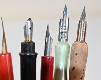 Puede incluir: Cinco plumas caligráficas con plumines metálicos y mangos variados. Los mangos son de color rojo, negro, rojo oscuro, verde pálido y corcho. Los plumines metálicos tienen textos grabados, incluyendo Falcon, con signos de desgaste.