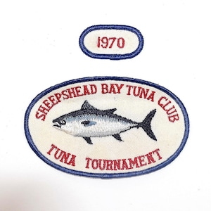 May include: Two embroidered patches with white backgrounds and blue borders. The larger oval patch features a tuna fish illustration and the red text "SHEEPSHEAD BAY TUNA CLUB TUNA TOURNAMENT". The smaller oval patch contains the red text "1970".