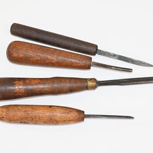 Könnte beinhalten: Eine Sammlung von vier Holzmeißeln mit abgenutzten Holzgriffen in verschiedenen Brauntönen. Jedes Werkzeug verfügt über einen Metallschaft und eine scharfe Klinge, die Gebrauchsspuren mit Kratzern auf den Holz- und Metalloberflächen aufweisen.