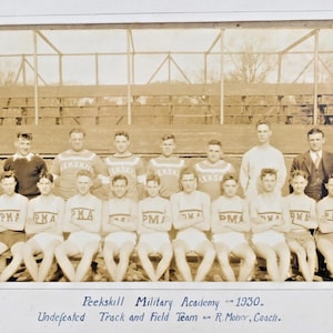 May include: A sepia photograph of the 1930 Peekskill Military Academy track and field team. The team, in athletic uniforms with "PMA" on the front, is seated in front of a wooden structure. The text at the bottom reads "Undefeated Track and Field Team."