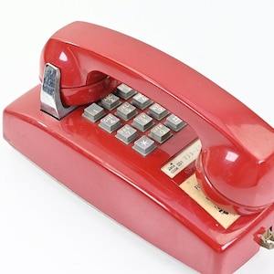 May include: A vintage red telephone with a handset and a keypad. The phone has a glossy finish and a silver metal cradle. The keypad features gray buttons with white lettering. A small label with text is visible on the base.