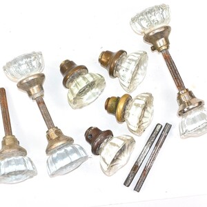 May include: A collection of vintage door knobs featuring clear, fluted glass handles attached to brass-toned metal bases. The set includes several knobs with attached metal spindles and two separate, thin, threaded metal rods for installation.