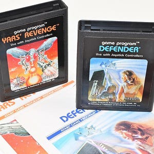 Può includere: Due cartucce rettangolari nere per videogiochi Atari 2600, etichettate Yars' Revenge e Defender, con etichette illustrate a colori e testo. Sotto le cartucce si trovano due manuali di istruzioni cartacei corrispondenti con la grafica del gioco.