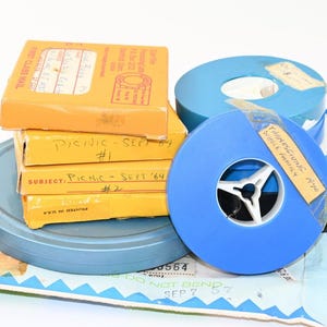 May include: A collection of vintage film reels and boxes. Three yellow boxes are stacked, labeled "Picnic - Sept '64 #1" and "#2". Two blue plastic film reels are present, one with a tape label reading "Thanksgiving 1970 Whole Family".