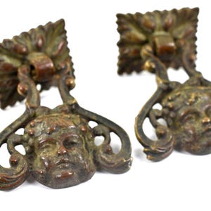 May include: Two matching bronze-toned metal drawer pulls featuring a square mounting plate with a leaf-like border and a swinging drop handle. The handle is shaped as a sculpted cherub face surrounded by ornate, curved metal scrollwork details.