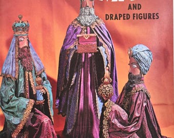 Op de afbeelding: Omslag van het tijdschrift "Papier Mache and Draped Figures". De afbeelding toont drie figuren in sierlijke gewaden en hoofddeksels. De figuren zijn in tinten paars, groen en blauw, met gouden details. De titel van het tijdschrift staat in grote witte letters.