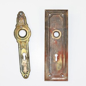 May include: Two antique door hardware pieces. One is a decorative, ornate brass door handle with a keyhole. The other is a rectangular brass door plate with a keyhole. Both pieces show signs of age and wear.