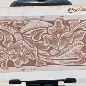 May include: A sepia-toned leathercraft pattern for a billfold, featuring floral and scroll designs. The design includes the text "BILLFOLD" and "Craftaid No. 2770". The letter "R" is prominently displayed. Measurements are indicated throughout the design.