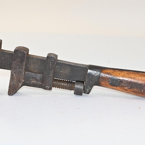 May include: A vintage adjustable wrench featuring a dark, weathered iron head and a sliding jaw mechanism. The tool has a threaded adjustment screw and a smooth, light brown wooden handle secured with two dark metal rivets on each side of the grip.