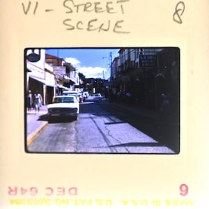 May include: A vintage 35mm film slide featuring a street scene with a white car parked on the left side. The slide mount has handwritten text reading VI - STREET SCENE and the number 8, with DEC 64R and MADE IN U.S.A. U.S. PAT. NO. 3,013,354 printed below.