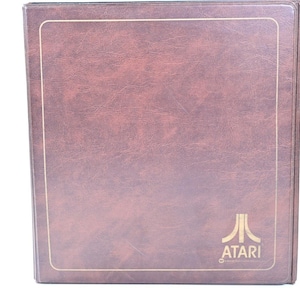 Puede incluir: Una carpeta rectangular de color marrón con un acabado de piel sintética texturizada. La portada presenta una fina línea de borde dorada y el logotipo y el texto de Atari en color dorado impresos en la esquina inferior derecha, sobre un fondo liso.