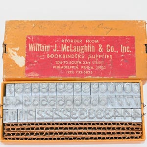 May include: A vintage orange cardboard box containing rows of silver metal letterpress type characters. The red label reads: REORDER FROM William J. McLaughtin & Co., Inc. BOOKBINDERS SUPPLIES 256-70 SOUTH 23rd STREET PHILADELPHIA, PENNA. 19103 (215) 732-5823.