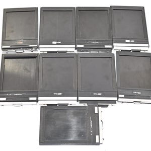 May include: A collection of nine black rectangular film holders for large format photography. Each unit features a dark central panel, a white bottom edge, and a metallic pull tab. The brand name Fidelity Elite is printed on several units.
