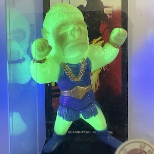 May include: A neon green gorilla figurine in a fighting stance, wearing a blue outfit with gold accents and a gold chain. The figurine is displayed in a clear case with Japanese text and French text in the background.