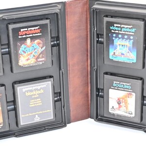 Può includere: Una custodia in plastica nera contenente otto cartucce di gioco Atari vintage. I titoli includono Backgammon, Superman, Video Pinball, Berzerk, Math Gran Prix, Blackjack, Bowling e un gioco Atari con etichetta bianca, tutti disposti in scomparti.