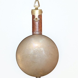 May include: A clock pendulum featuring a large, circular, metallic brass-toned disc attached to a vertical wooden rod. The top of the rod has a brass mounting bracket with a triangular loop, and the bottom of the disc has a small brass adjustment nut.
