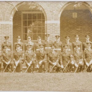 May include: A sepia photograph showing a group of uniformed individuals posing outdoors. The group is arranged in rows, with some kneeling and others standing. Each person holds a sword. The background features a brick building with arched openings.