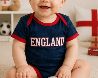 May include: A smiling baby wearing a navy blue onesie with red trim and the word "ENGLAND" in white. The baby is holding a soccer ball and is surrounded by England-themed decorations, including a teddy bear and flags.