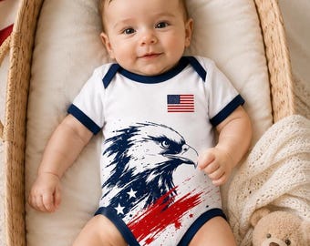 May include: A baby wearing a white onesie with a patriotic eagle design in blue, red, and white. The onesie has a small American flag and navy blue trim. The baby is lying in a wicker bassinet with a teddy bear.