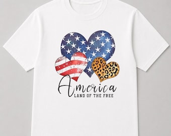 May include: White t-shirt featuring a design with three heart shapes. One heart has a pattern of the American flag, another has a blue starry sky pattern, and the third has a leopard print. The word "America" is written in cursive, with "LAND OF THE FREE" below.