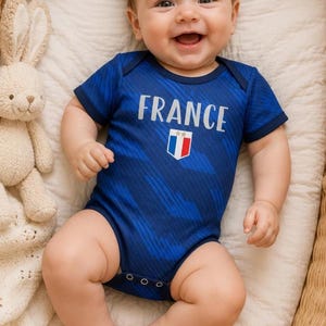 May include: A smiling baby wearing a blue bodysuit with the word "FRANCE" and a French flag emblem. The baby is lying in a wicker basket with a stuffed bunny and a small French flag nearby.