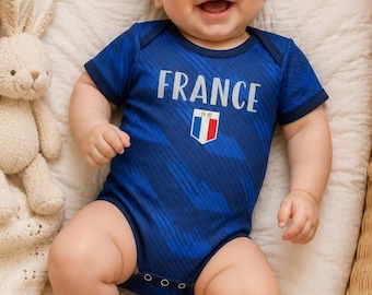May include: A smiling baby wearing a blue bodysuit with the word "FRANCE" and a French flag emblem. The baby is lying in a wicker basket with a stuffed bunny and a small French flag nearby.
