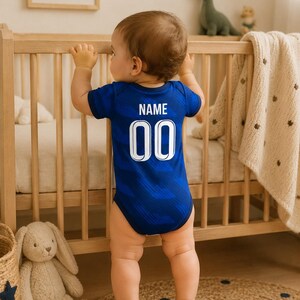 May include: A baby wearing a blue bodysuit with the word "NAME" and the number "00" printed on the back. The baby is standing in a nursery, holding onto a wooden crib. A stuffed bunny and a woven basket are visible.