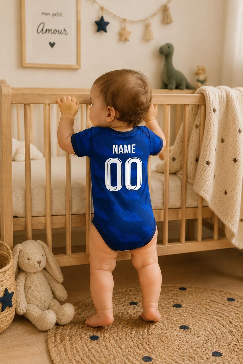 May include: A baby wearing a blue bodysuit with the word "NAME" and the number "00" printed on the back. The baby is standing in a nursery, holding onto a wooden crib. A stuffed bunny and a woven basket are visible.