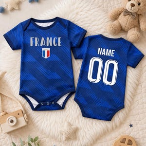 May include: Two blue baby onesies with the word "FRANCE" and a French flag graphic on the front. The back of one onesie has "NAME" and the number "00". The onesies are on a white fluffy surface.