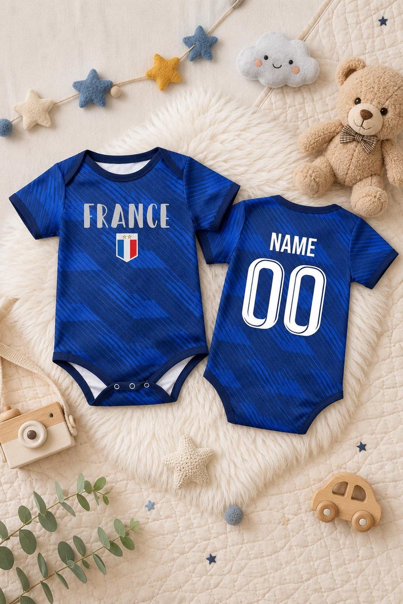 May include: Two blue baby onesies with the word "FRANCE" and a French flag graphic on the front. The back of one onesie has "NAME" and the number "00". The onesies are on a white fluffy surface.