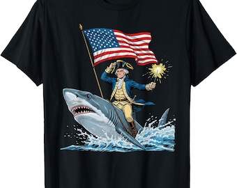 May include: Black t-shirt featuring a graphic of a man in colonial attire riding a shark, holding an American flag and a sparkler. The design is a patriotic and humorous illustration.