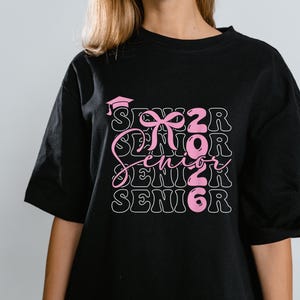 Puede incluir: Camiseta negra con un diseño rosa que dice "Senior 2026". El diseño incluye un gorro de graduación y un lazo. La palabra "Senior" se repite tres veces en un diseño apilado.