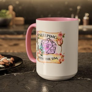 Puede incluir: Una taza grande de cerámica blanca con interior y asa rosas. La taza presenta el texto "SLEEPING WITH THE DM" con un gráfico de dados y detalles florales. La taza está sobre una superficie oscura.
