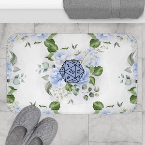 May include: A rectangular bath mat featuring a white background with a pattern of light blue flowers and green leaves. Centered on the mat is a light blue twenty-sided die graphic with black numbers and outlines, including the number 20 at the top.