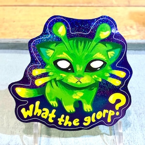 Puede incluir: Una pegatina de gato alienígena verde con ojos blancos grandes, antenas amarillas y un signo de interrogación. La pegatina tiene un fondo azul oscuro y morado con las palabras "what the glorp?" en amarillo.