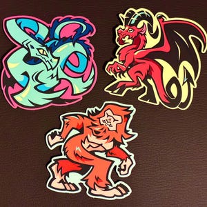 Può includere: Tre adesivi colorati raffiguranti creature fantasy. Uno è un serpente turchese con accenti rosa e blu. Un altro è un drago rosso con ali nere. Il terzo è una creatura arancione simile a Bigfoot.