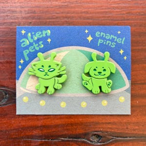 May include: Two lime green enamel pins shaped like a cat and a dog with antennae, displayed on a blue backing card featuring a green flying saucer illustration and the text alien pets enamel pins with yellow stars.