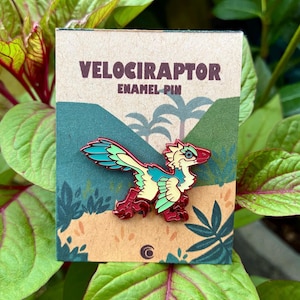 May include: A Velociraptor enamel pin attached to a rectangular cardboard backing card. The pin features a stylized dinosaur with cream, teal, and red feathers. The backing card displays the text VELOCIRAPTOR ENAMEL PIN above a jungle scene.