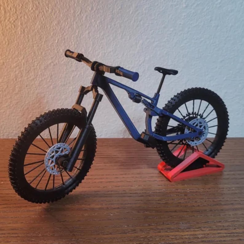 May include: A miniature blue mountain bike with black tires and components. The bike is displayed on a red stand, showcasing its design. The tires have a knobby tread pattern, and the bike has a suspension system.