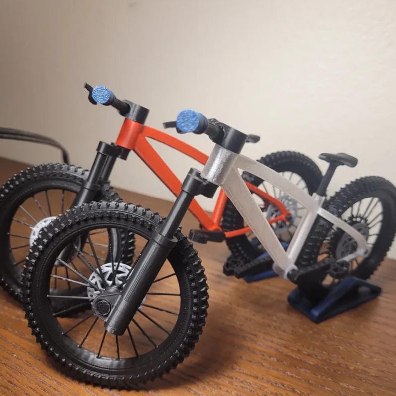 May include: Two miniature mountain bikes. One is red, the other is silver. Both have black tires, forks, and handlebars with blue grips. The bikes are displayed on a wooden surface.