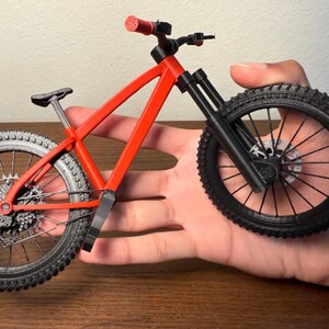 May include: A miniature red mountain bike with black tires and components. The bike has a detailed frame, suspension, and textured tires. The handlebars and seat are also black. The bike is held in a hand.
