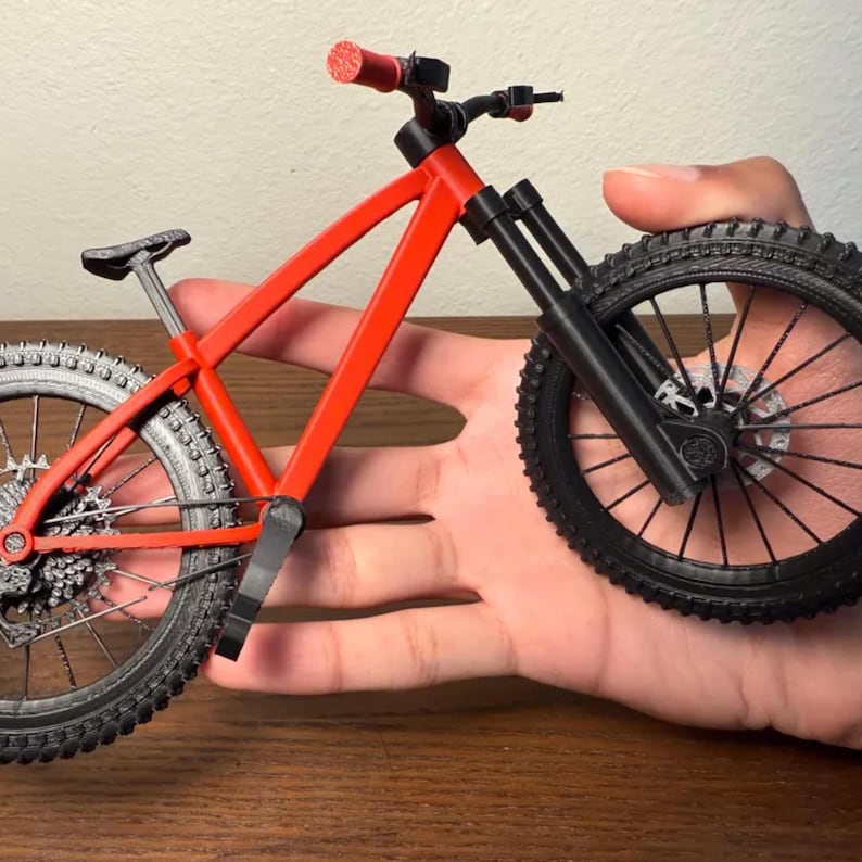 May include: A miniature red mountain bike with black tires and components. The bike has a detailed frame, suspension, and textured tires. The handlebars and seat are also black. The bike is held in a hand.