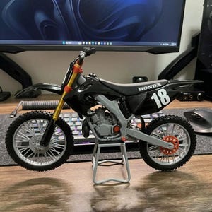 K&ouml;nnte beinhalten: Ein schwarzes und graues Honda Modellmotorrad mit der Nummer 18 auf der Seite. Das Motorrad hat goldene und orangefarbene Akzente und wird auf einem kleinen St&auml;nder ausgestellt. Die R&auml;der sind grau mit orangefarbenen Details.