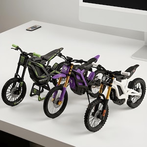 May include: Three miniature dirt bike models featuring detailed frames, knobby tires, and handlebars. The first is black and green with Monster branding, the second is purple with gold forks, and the third is white with the number 67 on the front plate.