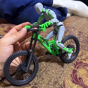 Könnte beinhalten: Ein grünes und schwarzes Mountainbike-Modell mit einer grauen, beweglichen Fahrerfigur. Das Fahrrad hat schwarze Reifen und einen grünen Rahmen. Der Fahrer befindet sich in Fahrposition, mit grauen und grünen Akzenten auf der Figur.