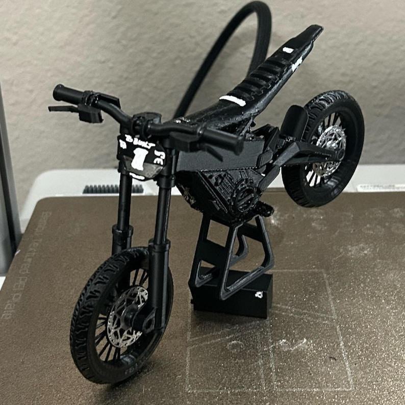 Puede incluir: Un modelo de motocicleta en miniatura negro con un dise&ntilde;o detallado. El modelo presenta un asiento texturizado, manillar y ruedas. La rueda delantera est&aacute; en el suelo, mientras que la rueda trasera est&aacute; levantada. El texto "SE" y el n&uacute;mero "1" son visibles en la parte delantera.