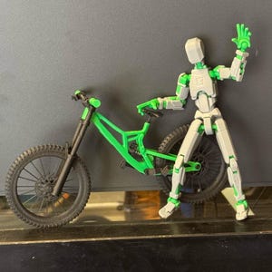 K&ouml;nnte beinhalten: Ein gr&uuml;nes und schwarzes Mountainbike mit detailliertem Design. Eine bewegliche Actionfigur in Wei&szlig; und Gr&uuml;n steht neben dem Fahrrad, mit einem Arm zum Gru&szlig; erhoben. Die Figur hat Gelenke.