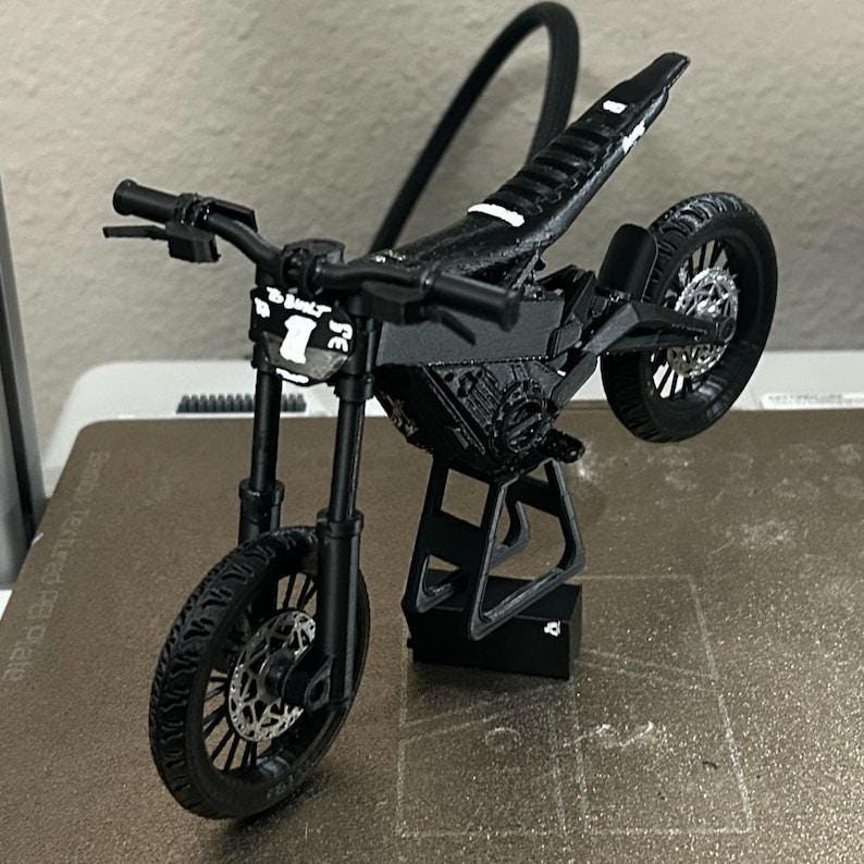 K&ouml;nnte beinhalten: Ein schwarzes Miniatur-Motorradmodell mit detailliertem Design. Das Modell wird von einem St&auml;nder gehalten, der das Vorderrad und den Lenker zeigt. Der Text "TO BE BUILT" und die Zahl "7" sind auf der Vorderseite sichtbar.
