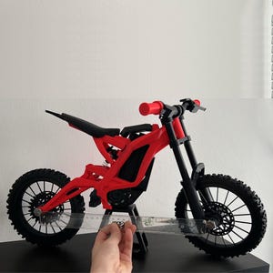 May include: A red and black miniature dirt bike model. The model has black tires, a black seat, and red handlebars. The bike is on a black surface. A ruler is held next to the bike to measure its size.